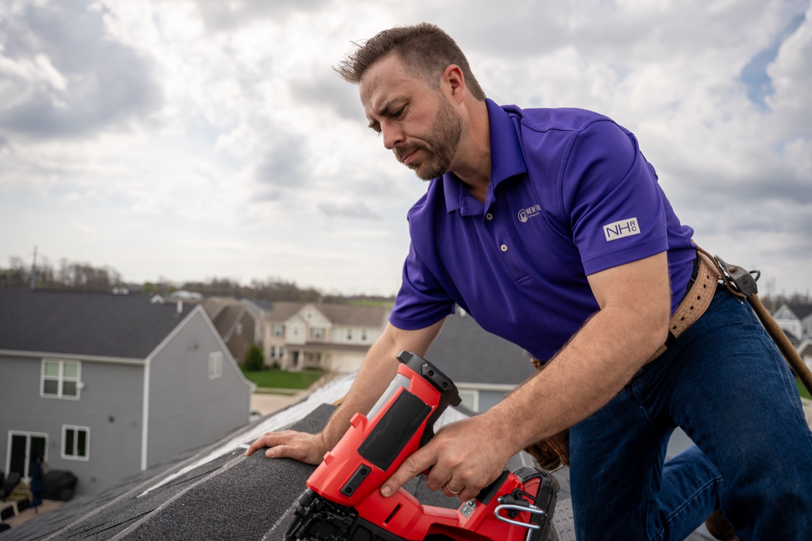 A roofer in a branded polo running a nail gun along a shingled roofline above a neighborhood